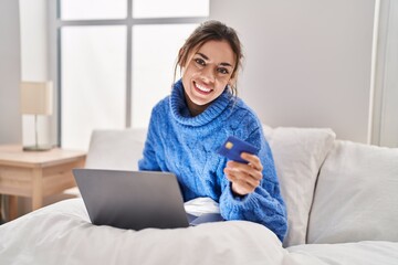 Young beautiful hispanic woman using laptop and credit card sitting on bed at bedroom