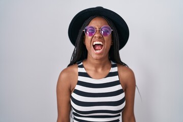 Young african american with braids wearing hat and sunglasses angry and mad screaming frustrated and furious, shouting with anger. rage and aggressive concept.