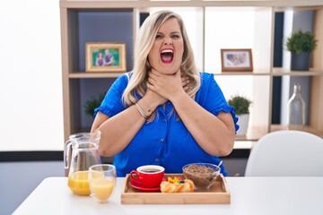 Obraz na płótnie Canvas Caucasian plus size woman eating breakfast at home shouting and suffocate because painful strangle. health problem. asphyxiate and suicide concept.