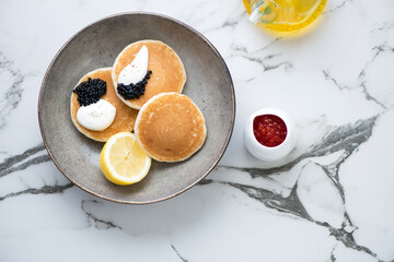 Plate of pancakes served with black caviar and sour-cream, above view on a white marble background, horizontal shot with space