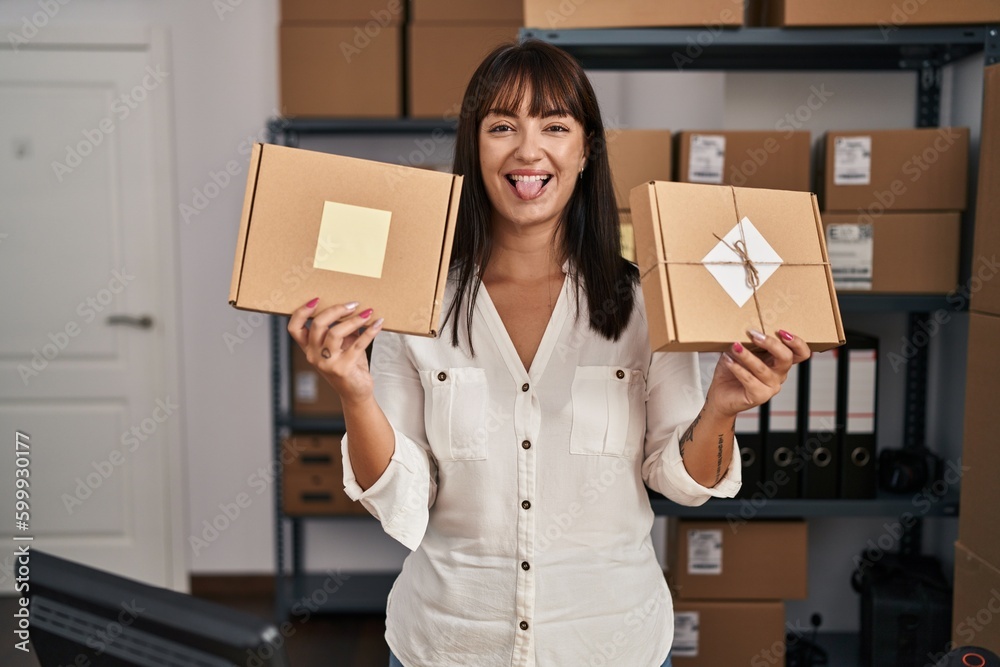 Sticker Young brunette woman working at small business ecommerce holding packages sticking tongue out happy with funny expression.