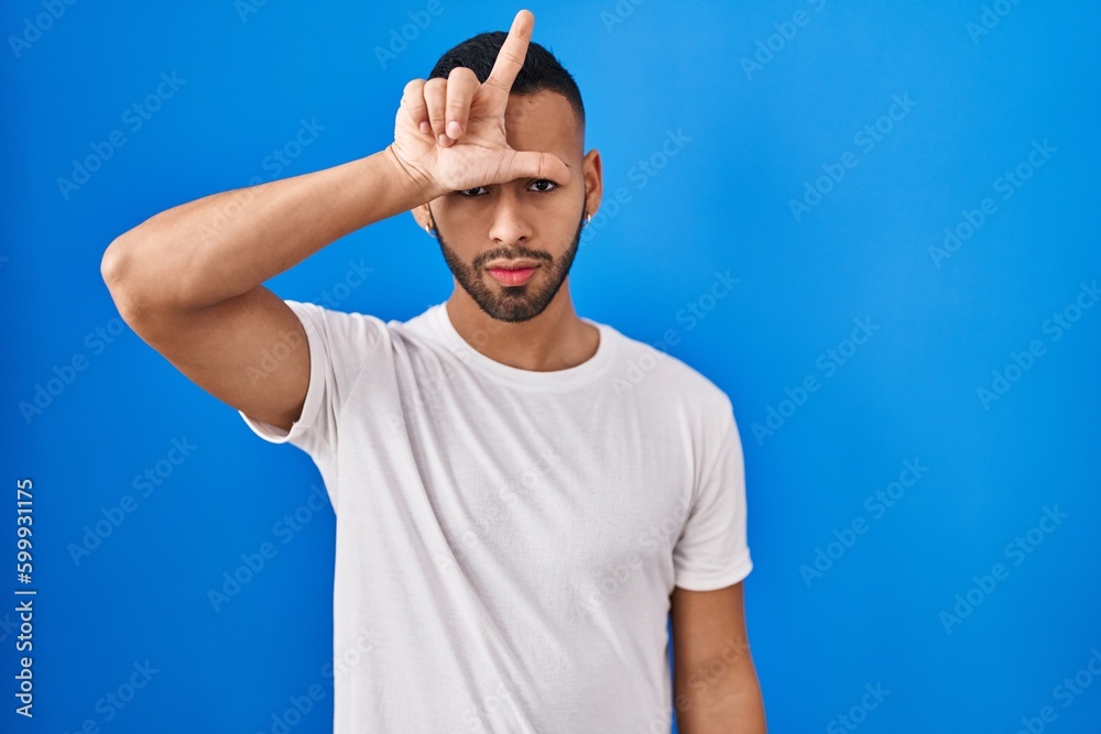 Poster young hispanic man standing over blue background making fun of people with fingers on forehead doing