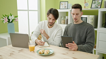 Two men couple having breakfast using touchpad and laptop writing on notebook at dinning room