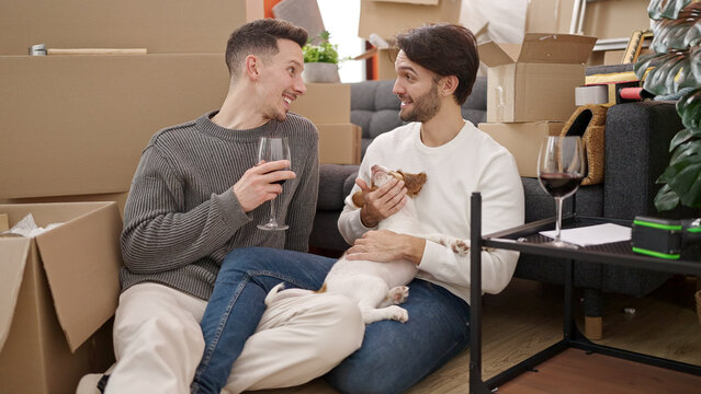 Two men couple drinking glass of wine sitting on floor with dog at new home
