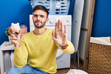 Hispanic man doing laundry holding piggy bank with open hand doing stop sign with serious and confident expression, defense gesture