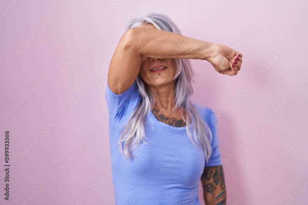 Wall mural middle age woman with tattoos standing over pink background covering eyes with arm smiling cheerful 