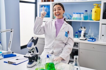 Young brunette woman working at scientist laboratory pointing finger up with successful idea. exited and happy. number one.