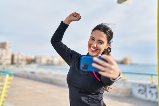 Young hispanic woman doing strong gesture making selfie by the smartphone at seaside