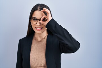 Young brunette woman standing over blue background doing ok gesture with hand smiling, eye looking through fingers with happy face.