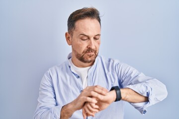 Middle age caucasian man standing over blue background checking the time on wrist watch, relaxed and confident