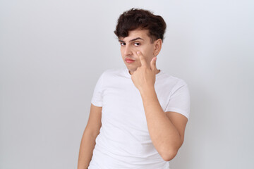 Young non binary man wearing casual white t shirt pointing to the eye watching you gesture, suspicious expression