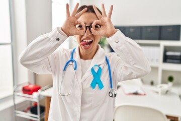 Young brunette doctor woman wearing stethoscope at the clinic doing ok gesture like binoculars sticking tongue out, eyes looking through fingers. crazy expression.