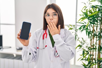 Young hispanic doctor woman showing smartphone screen covering mouth with hand, shocked and afraid...