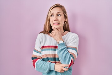 Young blonde woman standing over pink background thinking worried about a question, concerned and nervous with hand on chin