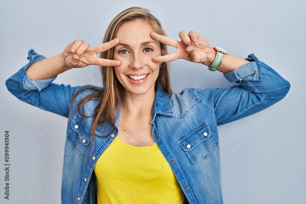 Sticker Young blonde woman standing over blue background doing peace symbol with fingers over face, smiling cheerful showing victory