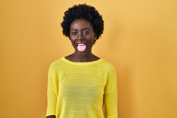 African young woman standing over yellow studio sticking tongue out happy with funny expression. emotion concept.