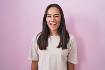 Fototapeta premium Young hispanic woman standing over pink background winking looking at the camera with sexy expression, cheerful and happy face.