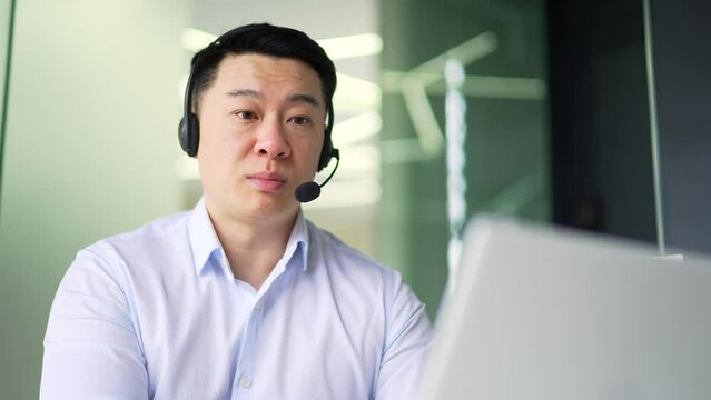 Corporate operator, asian man who works in customer support speaks on a video call using a laptop. Call center agent in wireless headset helping customer with complaints in office Online consultation