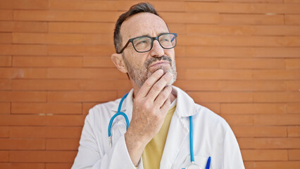 Middle age man doctor standing with doubt expression thinking over isolated brick background