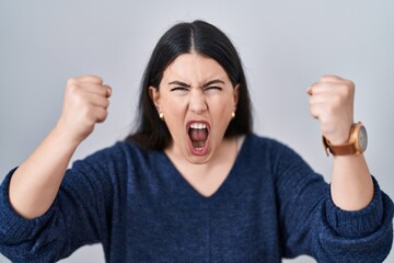 Young brunette woman standing over isolated background angry and mad raising fists frustrated and furious while shouting with anger. rage and aggressive concept.