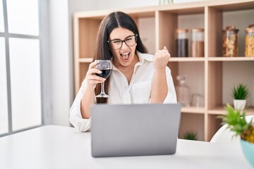 Young brunette woman doing video call with laptop drinking a glass of wine screaming proud, celebrating victory and success very excited with raised arm