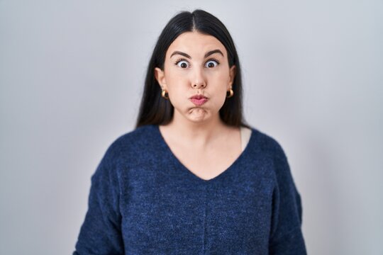 Young brunette woman standing over isolated background puffing cheeks with funny face. mouth inflated with air, crazy expression.