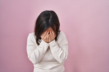 Woman with down syndrome standing over pink background with sad expression covering face with hands...