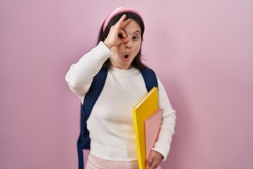 Woman with down syndrome wearing student backpack and holding books doing ok gesture shocked with surprised face, eye looking through fingers. unbelieving expression.