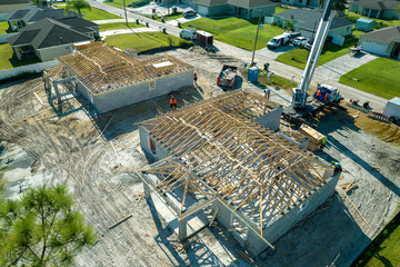 Aerial view of lifting crane and builders working on unfinished residential house with wooden roof frame structure under construction in Florida suburban area
