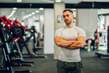 A confident muscular sportsman is posing in a gym with arms crossed.