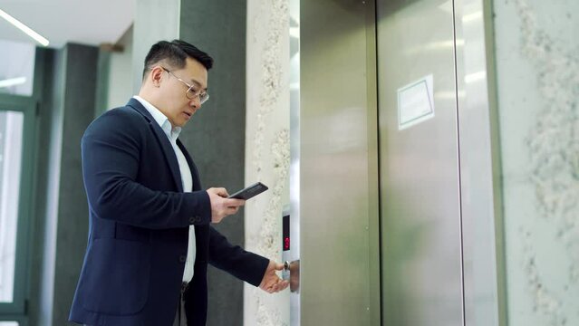 Asian businessman calling the elevator using a smartphone while browsing in a modern office building. Thoughtful entrepreneur in glasses looking at the phone while the lift door opens and goes inside