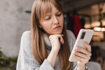 Confused puzzled blonde bang woman in casual clothes, sits in cafe, holds a smartphone in her hand, looks questioningly at the mobile phone, spreading her arm around. Girl getting surprising bad news.