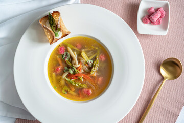 Top view of a vegetables soup with asparagus, mushroom and pink (beetroot) gnocchi in a white plate with bread variation. Golden spoon on the pink cloth. Vegetarian food. Gourmet restaurant.