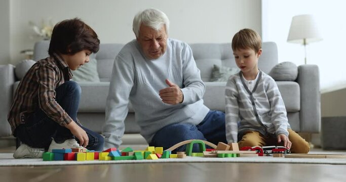 Older grandpa watching two grandkid boys, playing building blocks, constructing toy road on heating floor. Brother kids and grandfather enjoying learning activity at home, family leisure
