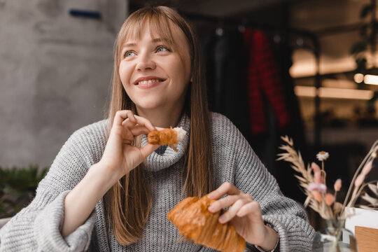 Blonde happy woman with bang having a relaxing moment at cafe breaking her croissant at small piece and eat it. Attractive caucasian girl wear grey sweater, enjoying a fragrant croissant, lunch time.