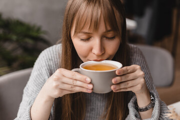 Happy young woman drinking a cup of tea in the morning. Dreaming girl sitting in restaurant with cup of hot tea enjoying with closed eyes. Pretty woman wearing grey sweater in cafe. Good day, resting.