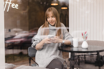 Young cute woman using phone sits in cafe at table with smartphone, answering texts, phone call, letters, posting photos under windows glass. Woman look dreaming, think about answer, wear grey sweater