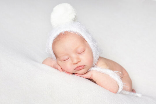 a newborn baby in a white hat with a pom-pom sleeps on a white bedspread over white background with copy space. beautiful sleeping baby