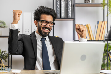 Indian businessman working on laptop at home office desk typing browsing say Wow yes found out great big lottery jackpot win, good job news, celebrating. Manager freelancer arab man
