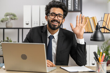 Hello. Indian business man working on laptop computer smiling friendly at camera and waving hands gesturing hi, greeting or goodbye, welcoming with hospitable expression at home office workplace desk