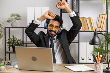 Happy relaxed overjoyed indian businessman working on laptop computer at home office wearing headphones listening favorite energetic disco music and dancing. Freelancer man relaxing, taking a break