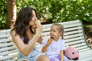 Mother and daughter having snack on bench after skating inline roller in public park together....