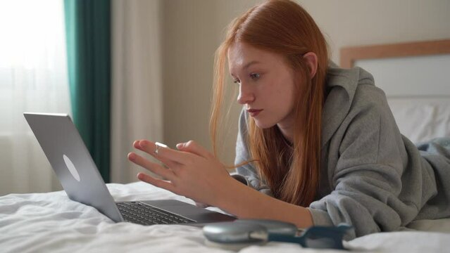 Young woman talking on smartphone while using laptop at home on bed. Teenage girl speaking with friends mobile phone working computer, studying typing, online shopping, watching video, reels