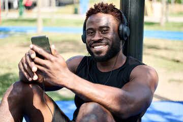 Happy fit sporty young black man sitting in workout park using mobile phone apps listening music. Strong African ethnic guy bodybuilder wearing headphones holding smartphone outdoors.
