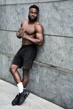 Fit strong shirtless young African man standing at urban wall. Sporty muscular black guy bodybuilder wearing black sport shorts resting after sport street workout training outdoor, vertical.