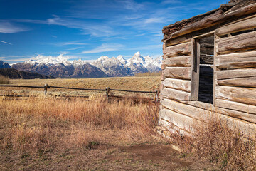 The side of a deserted homestead cabin, fence, autumn meadow and snow-capped Grand Teton Mountain Range in Grand Teton National Park Wyoming.