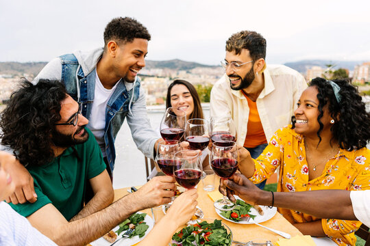 Multiracial group of young friends enjoying bbq dinner party on rooftop. Smiling millennial people celebrating together sitting on terrace table toasting red wine enjoying good moments in summer