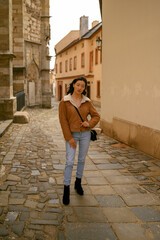 Young Asian model in fashionable clothing posing on a historic street in a European city. Beautiful cityscape and landmarks in the background. Ideal for travel, tourism, and fashion-related themes.