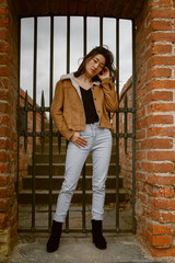 A young Asian model in stylish clothing poses confidently while leaning against an old gate with bars. The brick and urban setting.