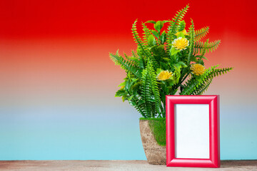 Top view of red empty picture frame standing on table and beautiful flower pot on blue red mix colors background with free space